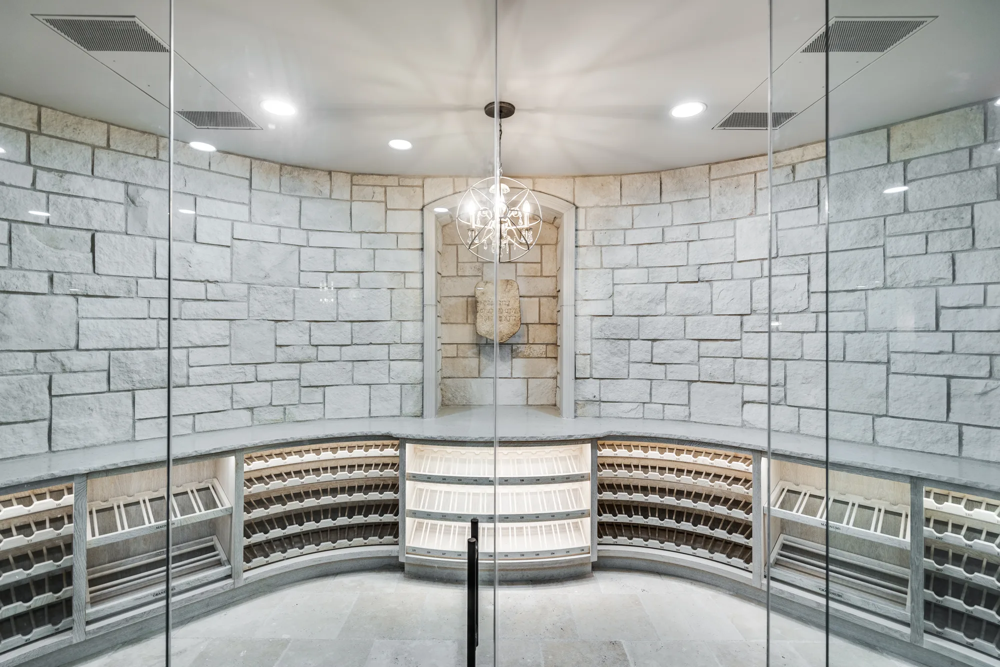 Wine room behind full-height glass, walled in cut Jerusalem stone with curved oak racking and a carved stone set into an arched niche