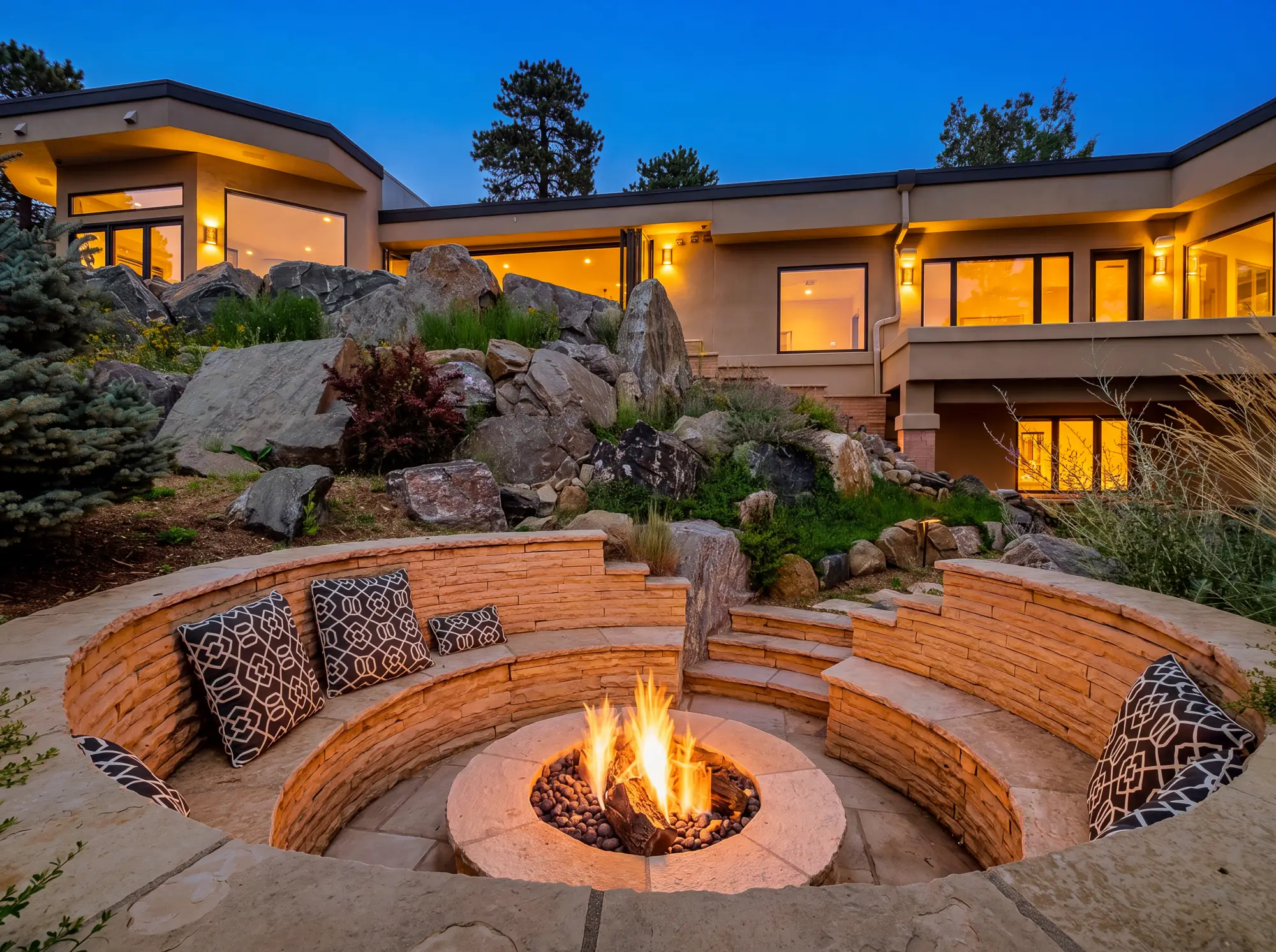 The fire pit lit at dusk with the house glowing behind the boulders.
