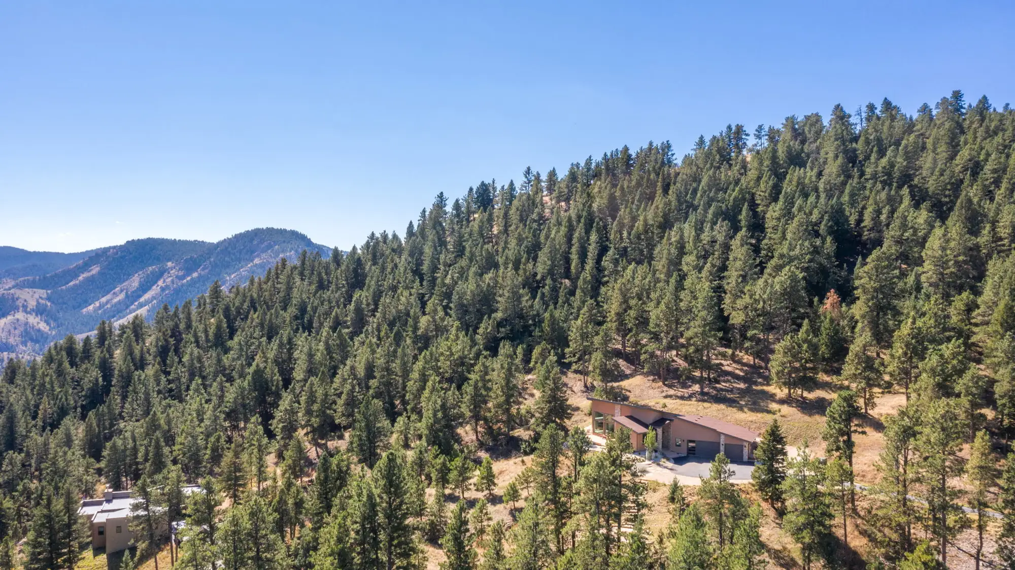 Wide aerial showing the house small against the forested canyon wall, with the range folding away behind