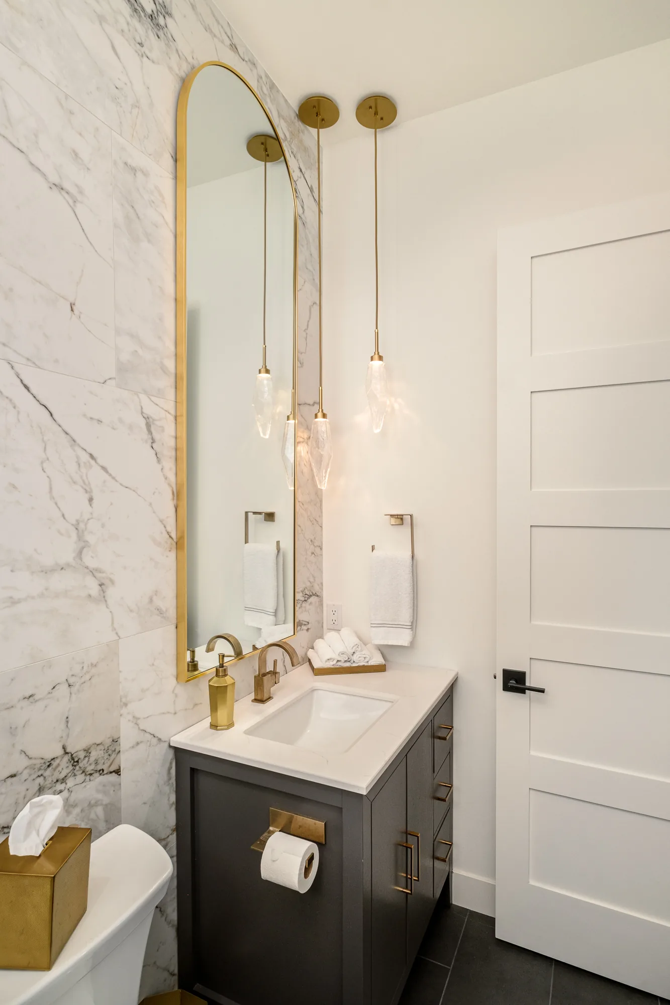 Guest bathroom with a white marble slab wall, tall arched gold mirror, quartz pendant lights and a charcoal vanity.