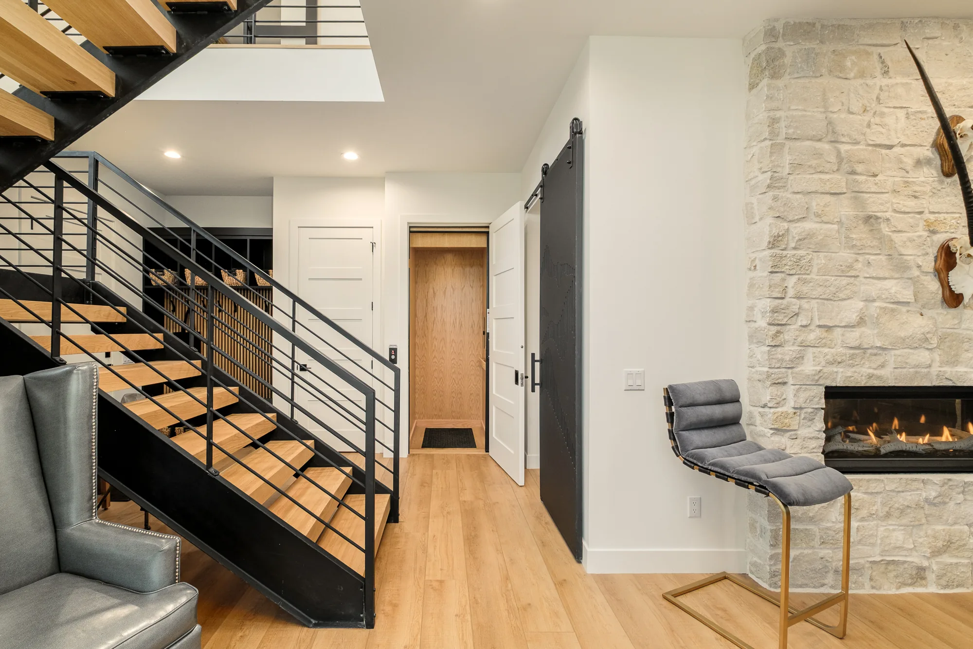 View from the lower level of an open-tread oak and black steel staircase beside a white stone fireplace and a black barn door.