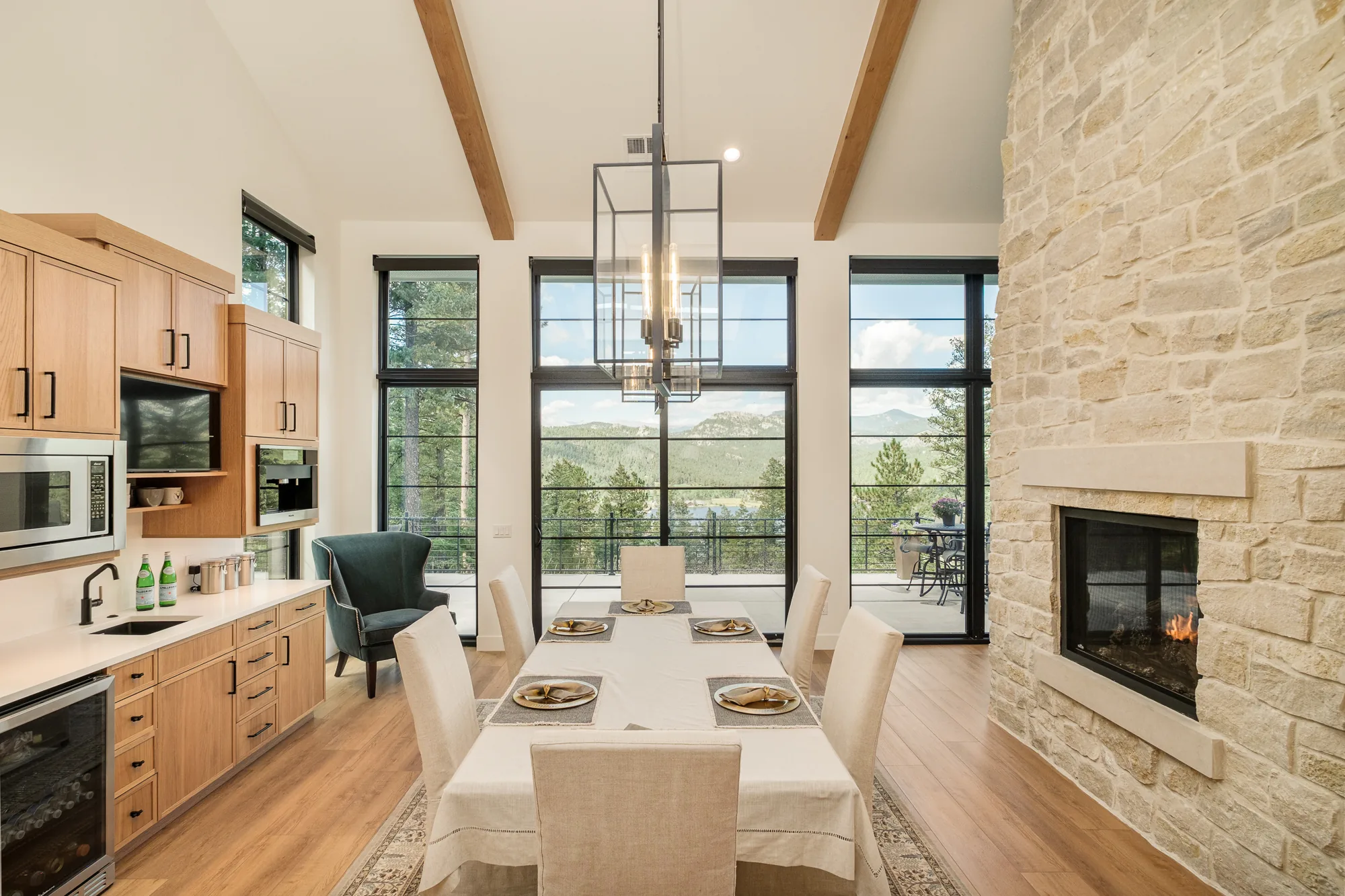 Dining area with a long table, black lantern chandelier and a two-sided stone fireplace, with mountain and lake views through tall windows.