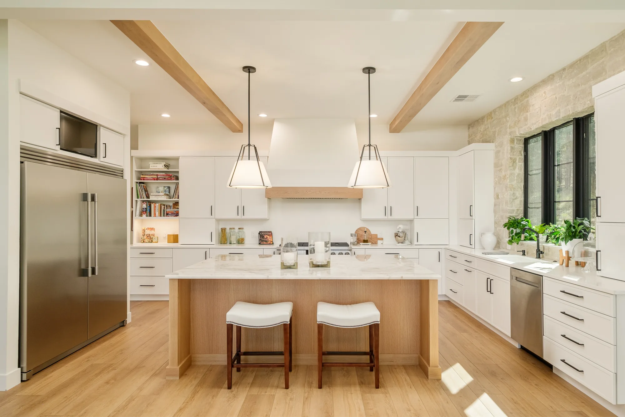 White kitchen with oak ceiling beams, a marble-topped white oak island, twin fabric pendants and a stone accent wall.