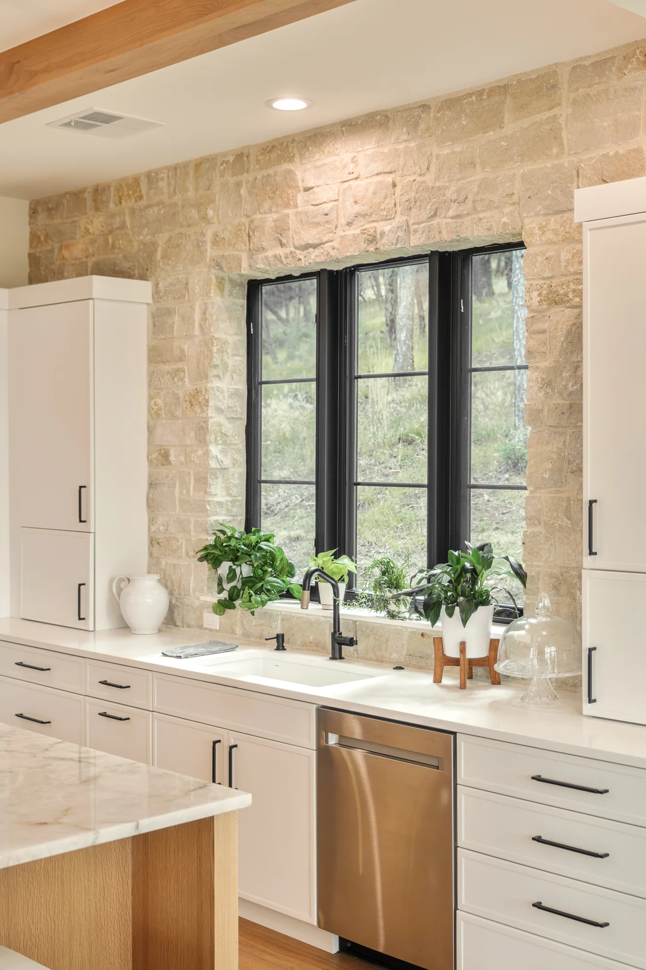 Prep sink set into a quarried stone wall beneath black steel windows, with herbs on the sill.