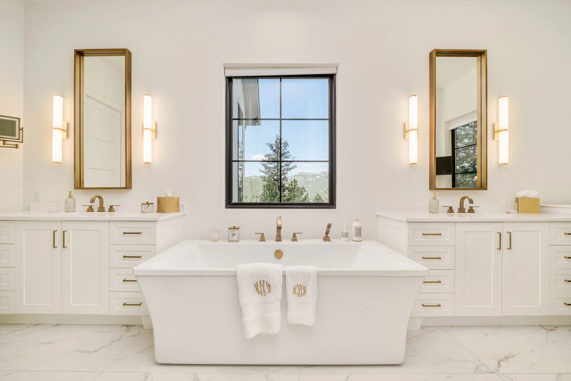 Primary bath with twin vanities, brass mirrors and sconces, and a freestanding tub beneath a window framing a pine and the range.