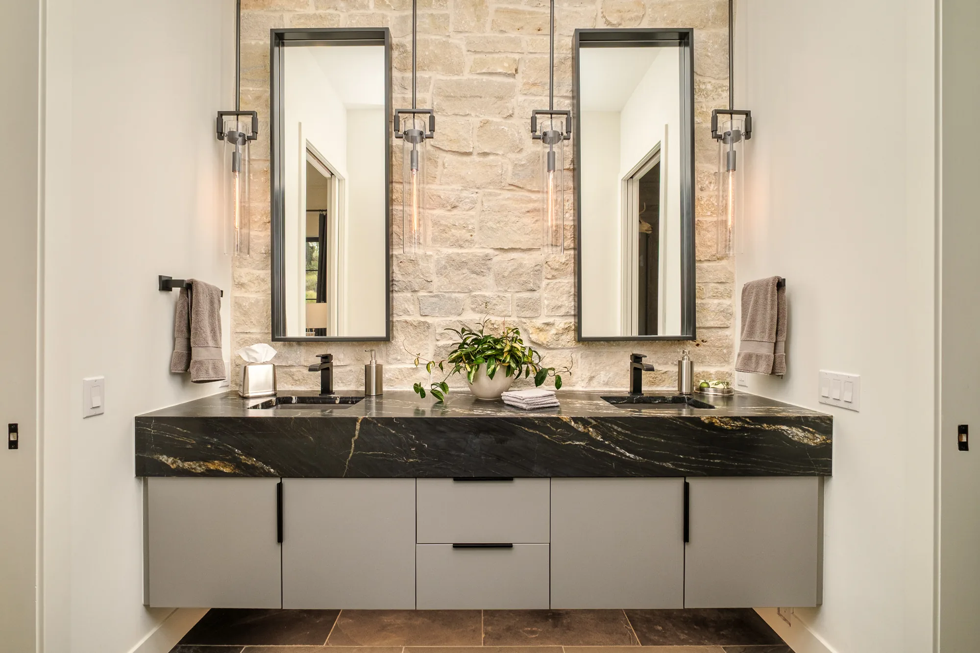 Floating double vanity with a thick black granite top, steel-framed mirrors and sconces against a cut stone wall.