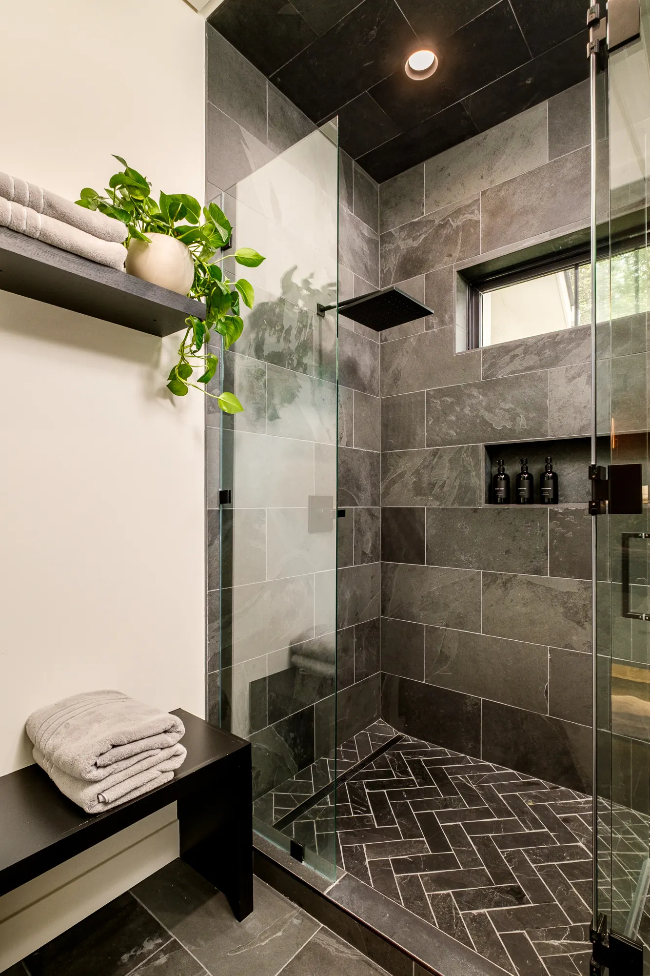 Walk-in shower clad in charcoal slate with a herringbone floor, glass enclosure and a blackened bench.