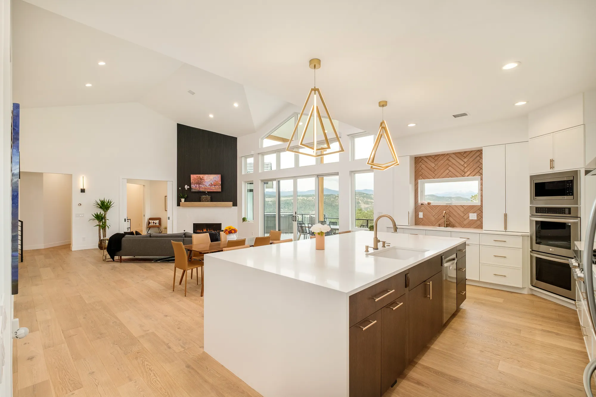 Kitchen with a long white island, gold geometric pendants and a black fireplace wall beyond